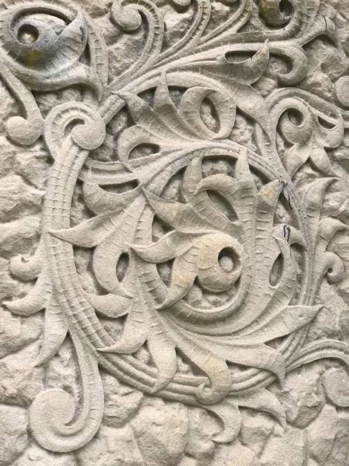 Intricate stone carving featuring swirling patterns and floral motifs on a textured surface.