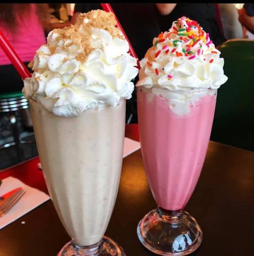 Two tall milkshakes in glasses: one beige with whipped cream and crumbs, the other pink topped with whipped cream and sprinkles.