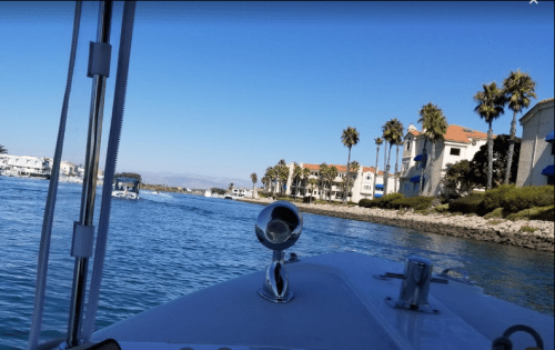 View from a boat on a calm waterway, with palm trees and waterfront buildings lining the shore under a clear blue sky.