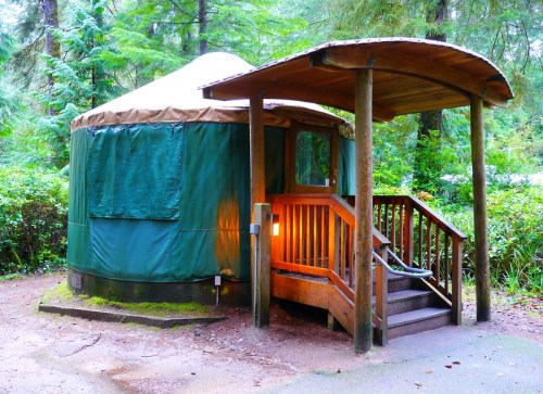 A cozy yurt with a wooden deck, surrounded by lush greenery in a forested area.