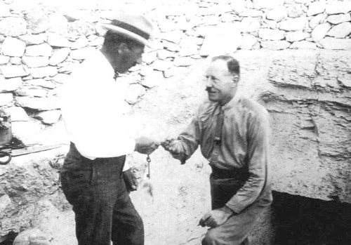 Two men shake hands in a rocky outdoor setting, one wearing a hat and the other in work attire.