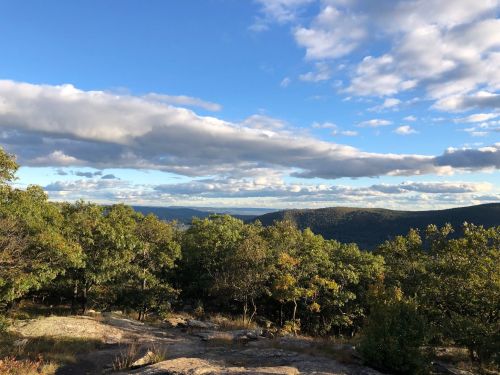 A scenic view of rolling hills and trees under a partly cloudy sky, with distant mountains visible on the horizon.