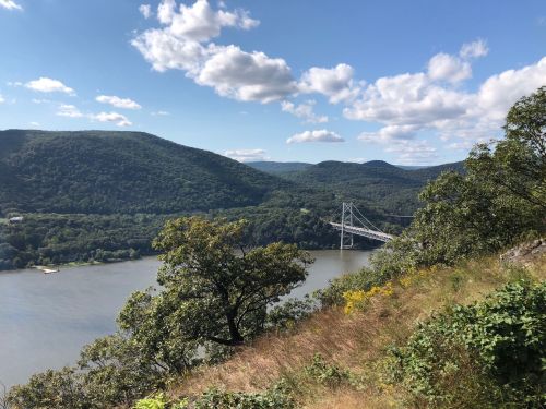 A scenic view of a river surrounded by green hills and a bridge under a blue sky with fluffy clouds.