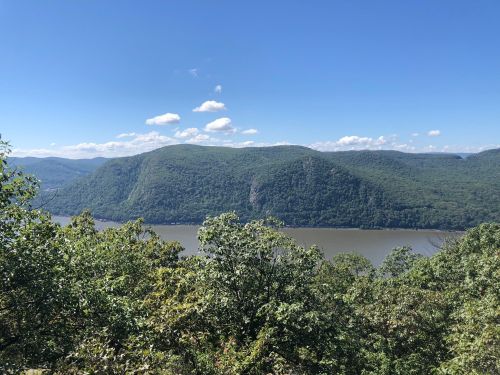 A scenic view of a river surrounded by lush green hills under a clear blue sky with scattered clouds.