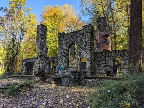 Ruins of a stone structure surrounded by colorful autumn trees and fallen leaves. Two people explore the site.