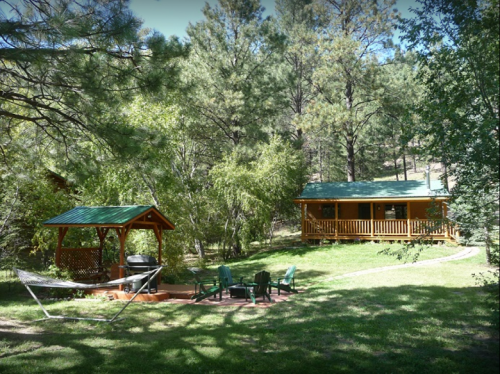 A cozy cabin surrounded by trees, with a gazebo, fire pit, and green lawn in a serene outdoor setting.
