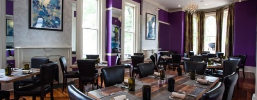 Elegant restaurant interior with purple walls, wooden tables, and black chairs, featuring large windows and stylish decor.