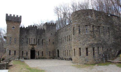 A stone castle with towers and large windows, surrounded by trees and a gravel pathway.