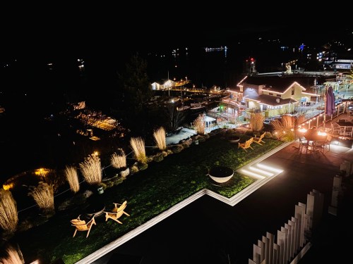 A nighttime view of a waterfront area with illuminated buildings, boats, and a landscaped terrace featuring chairs.