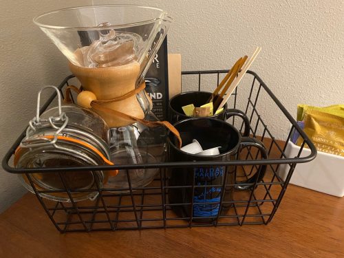A black wire basket containing a coffee maker, mugs, sugar packets, and jars on a wooden surface.