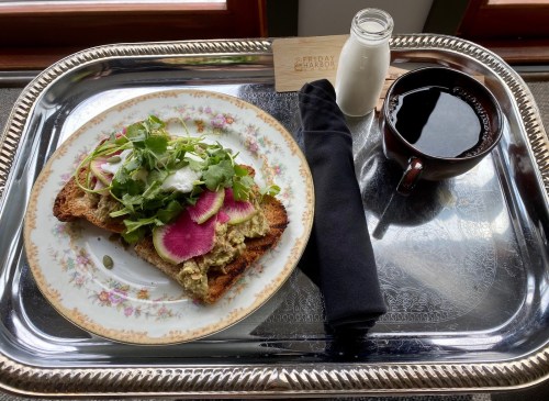 A tray with toast topped with hummus, greens, and radish, alongside a cup of coffee and a small bottle of cream.