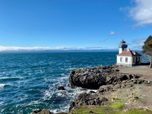 A lighthouse stands on rocky shore, overlooking a vibrant blue ocean under a clear sky with scattered clouds.
