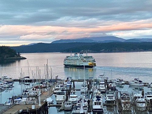 A ferry navigates through calm waters near a marina, with mountains and a colorful sky in the background.
