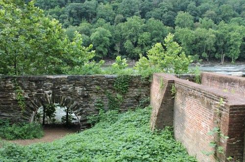 A stone archway surrounded by greenery, leading to a river, with remnants of brick walls in the foreground.