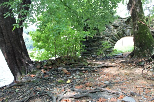 A stone archway partially hidden by trees and greenery, with roots and scattered rocks along the riverbank.