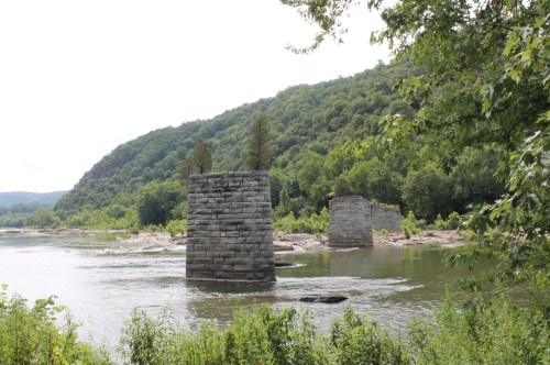Ruins of stone pillars rise from a river, surrounded by lush green trees and hills under a cloudy sky.