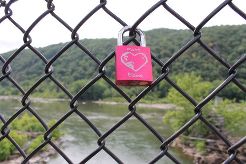 A pink padlock with names "Matt" and "Elaina" hangs on a chain-link fence overlooking a river and green hills.