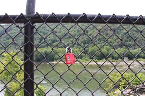 A red padlock hangs on a black chain-link fence overlooking a river and green hills in the background.