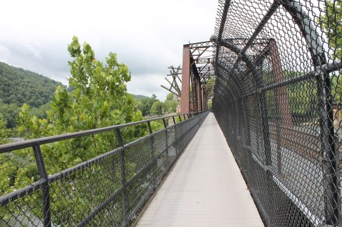 A walkway on a metal bridge surrounded by greenery and mountains under a cloudy sky.