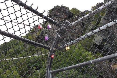 A close-up of a chain-link fence with colorful locks attached, overlooking a rocky landscape and greenery.
