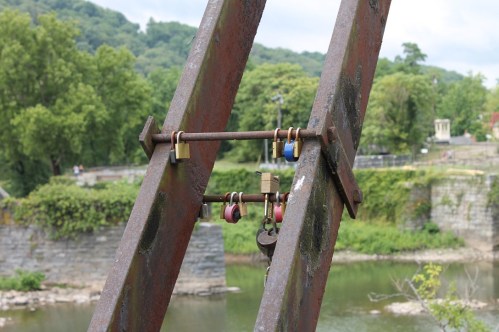 Rusty metal structure with colorful padlocks attached, overlooking a river and green trees in the background.