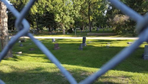 A view of a cemetery through a chain-link fence, featuring gravestones and green grass under a clear sky.