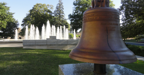 A bronze bell in the foreground with a fountain and trees in the background on a sunny day.
