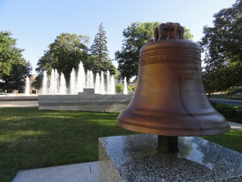 A large bronze bell on a stone pedestal, with a fountain and trees in the background under a clear blue sky.