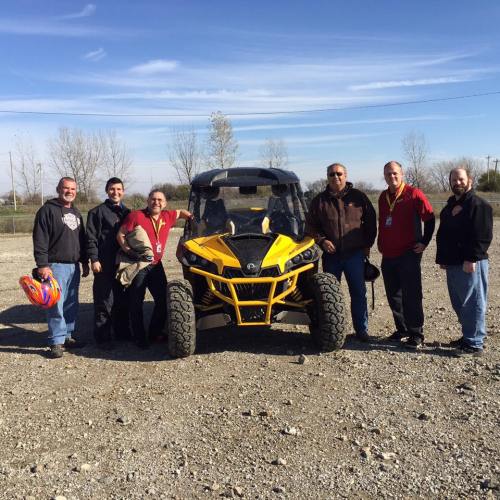 A group of six people stands beside a yellow off-road vehicle on a gravel surface under a clear blue sky.