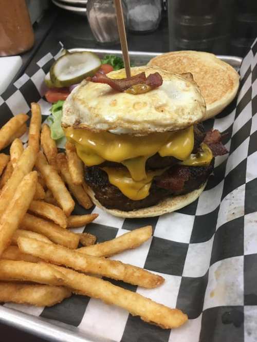 A double cheeseburger with bacon and a fried egg, served with fries and pickles on a checkered tray.