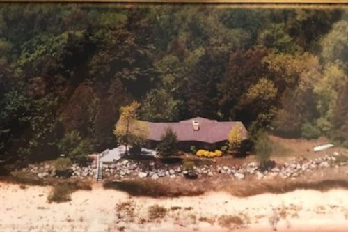 Aerial view of a house surrounded by trees, near a sandy beach and rocky shoreline.