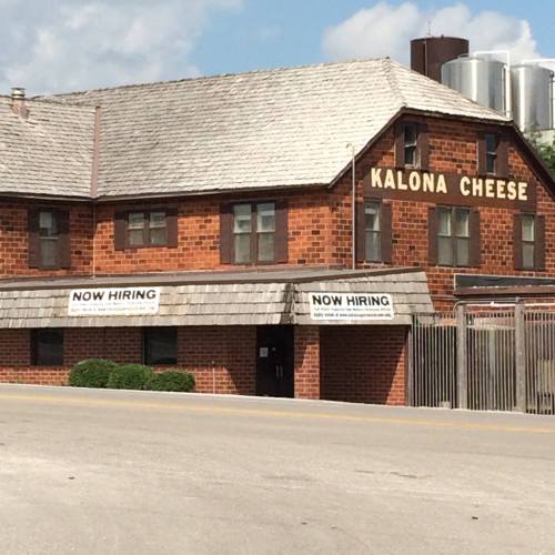 A brick building with a sign reading "KALONA CHEESE" and a "NOW HIRING" notice on the front.
