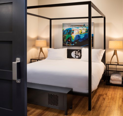A modern bedroom featuring a four-poster bed, two lamps, and a colorful artwork on the wall. Warm wooden floors.