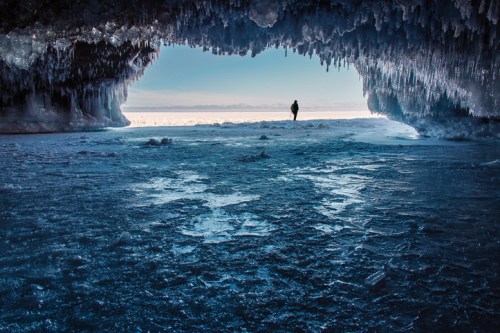 A person stands in an icy cave, framed by icicles, with a frozen lake visible in the background.