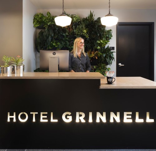 A smiling receptionist stands behind a modern hotel front desk with greenery in the background and a computer setup.