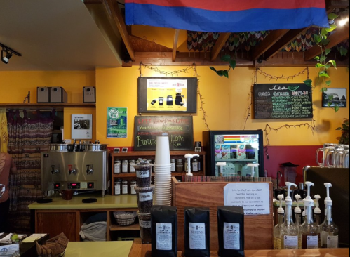 A cozy coffee shop interior with a counter, coffee equipment, and a colorful wall adorned with a flag and decorations.