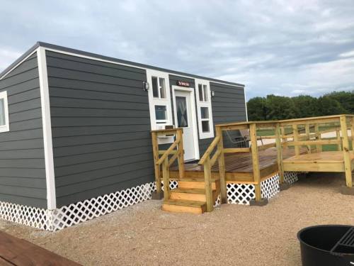 A small, modern cabin with a wooden deck, set in a grassy area under a cloudy sky.