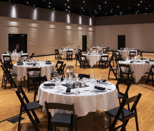 Elegant dining setup with round tables, white tablecloths, and black chairs in a spacious, well-lit hall.