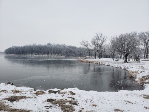 A serene winter landscape featuring a partially frozen lake, snow-covered banks, and bare trees under a gray sky.