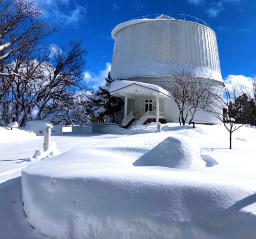 A snow-covered landscape featuring a large white observatory and trees under a bright blue sky.