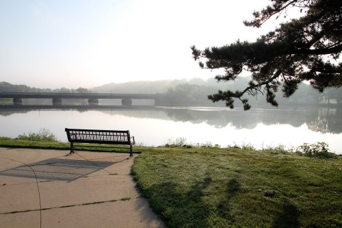 A serene riverside scene at dawn, featuring a bench, calm water, and a bridge in the background.