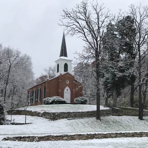 A brick church with a tall steeple surrounded by snow-covered trees and a grassy area.