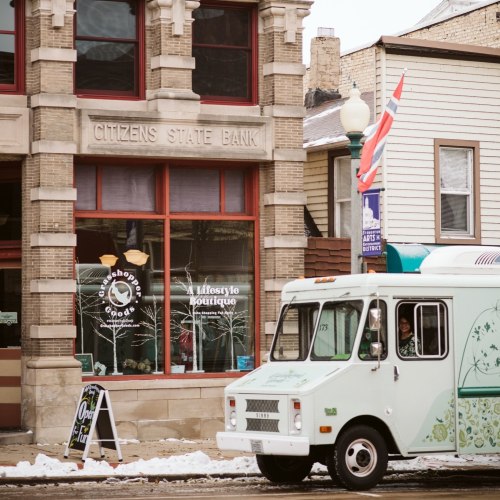 A vintage ice cream truck parked in front of a bank and boutique on a snowy street.