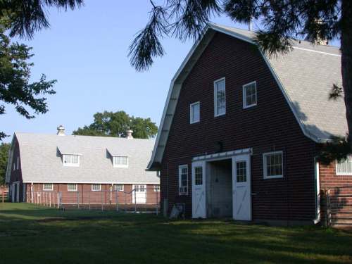 Two red brick barns with gray roofs, surrounded by green grass and trees under a clear blue sky.
