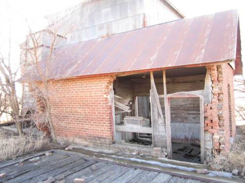 An old, weathered brick building with a damaged wall and a rusty metal roof, surrounded by overgrown vegetation.