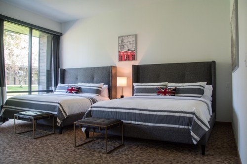 A modern hotel room featuring two beds with gray upholstery, striped bedding, and a view of a garden outside.