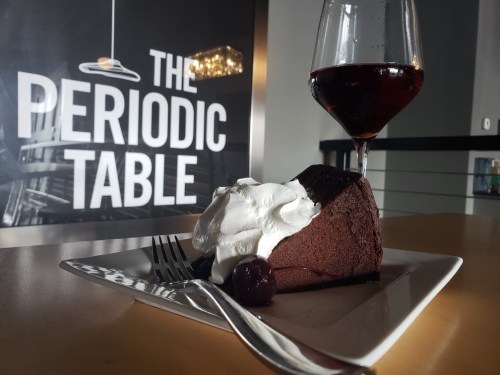 A slice of chocolate cake topped with whipped cream and a cherry, next to a glass of red wine, against a stylish backdrop.