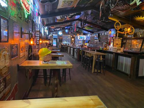 A rustic bar interior with wooden tables, neon signs, and a yellow hard hat on a table, creating a lively atmosphere.