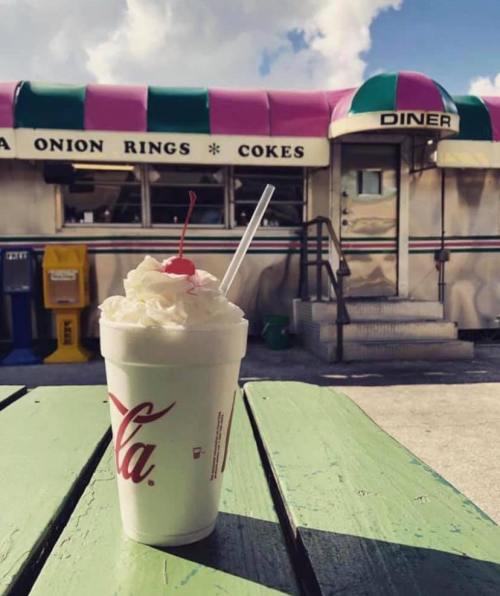 A milkshake topped with whipped cream and a cherry sits on a green table in front of a colorful diner.