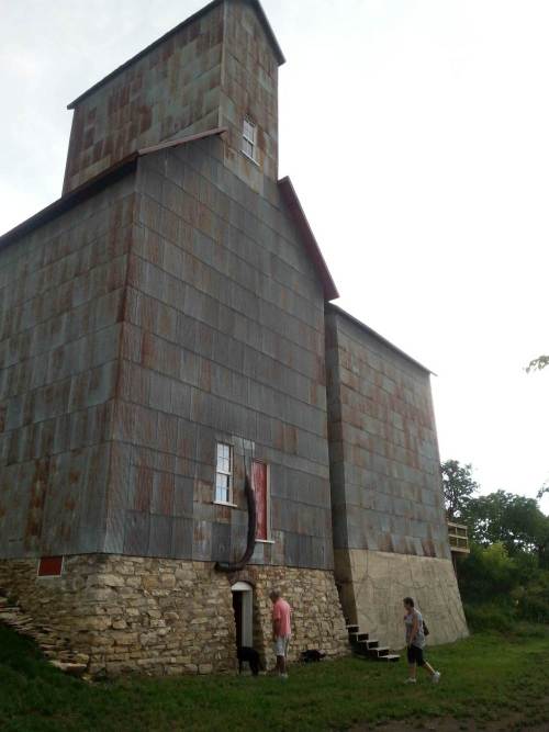 A tall, rustic building with a metal exterior, two people and two dogs are near the entrance on a grassy area.